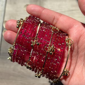 Könnte beinhalten: Ein Stapel roter Armreifen mit goldenen Akzenten. Die Armreifen sind tiefrot mit Goldflitter und durch goldene Ketten mit kleinen goldenen Blumenanhängern verbunden. Die Armreifen werden in einer Hand gehalten.