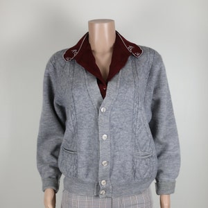 May include: A grey cable knit cardigan with a v-neck and button fastening. The cardigan has two pockets and is worn over a burgundy collared shirt with a white embroidered trim.