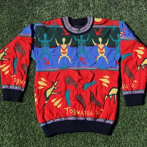 Pu&ograve; includere: Un maglione colorato con girocollo e maniche lunghe. Il maglione presenta una base rossa con dettagli blu, verdi e gialli. Presenta un motivo di omini stilizzati, frecce e forme astratte. I polsini e lo scollo sono blu scuro.