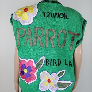 Puede incluir: Chaleco de punto verde sin mangas con las palabras "TROPICAL PARROT BIRD LA" bordadas en la espalda. El chaleco tiene aplicaciones de flores de colores en la parte delantera.