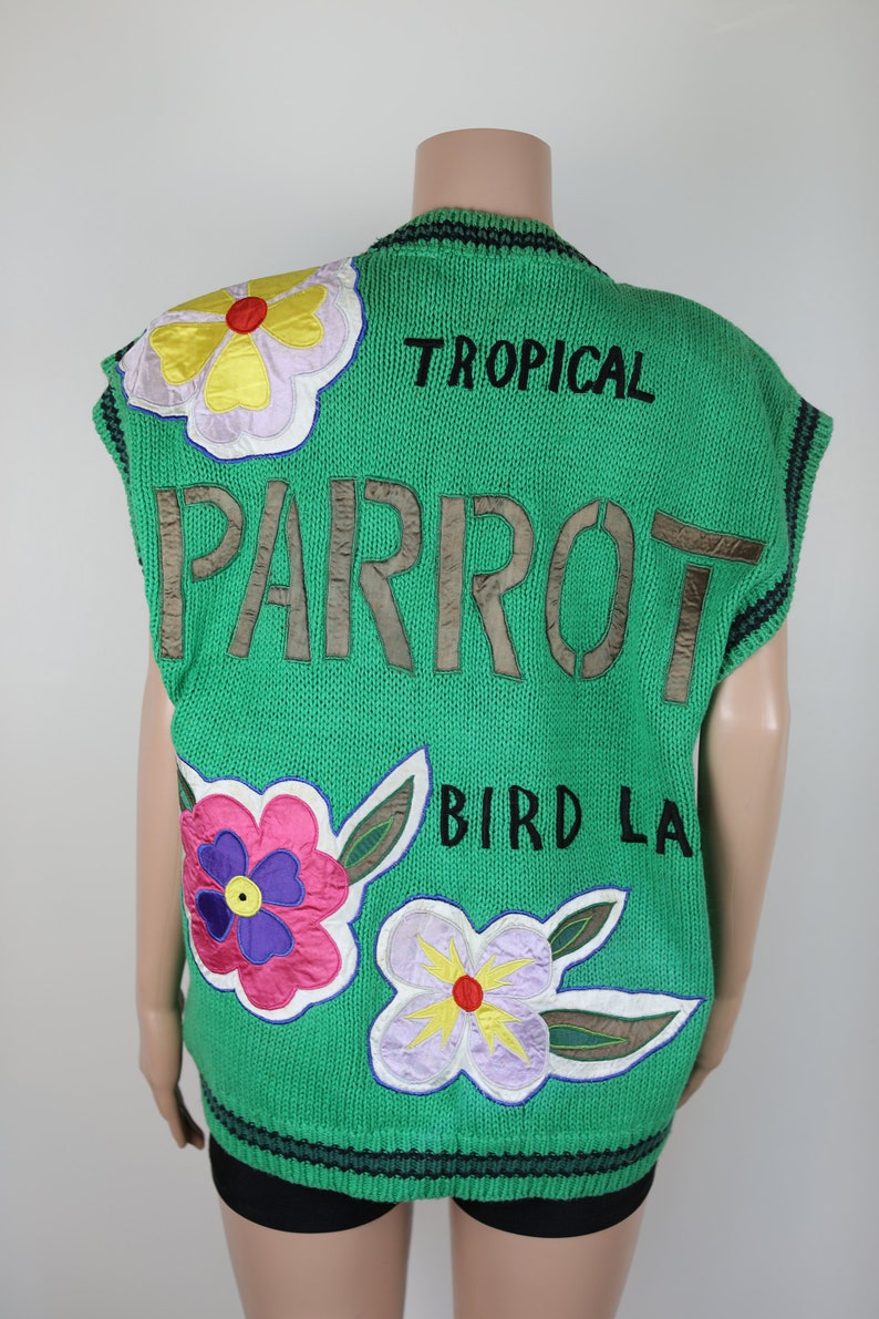 Puede incluir: Chaleco de punto verde sin mangas con las palabras "TROPICAL PARROT BIRD LA" bordadas en la espalda. El chaleco tiene aplicaciones de flores de colores en la parte delantera.