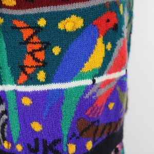 May include: A colorful knitted sweater with a parrot design. The sweater features a variety of colors, including red, blue, green, yellow, and orange. The parrot is depicted in a bright red, blue, and yellow color scheme. The sweater has a black background with a white stripe.