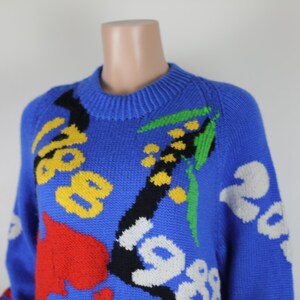 May include: A blue knitted jumper with a colourful abstract design. The design features yellow, red, green, black and white accents.