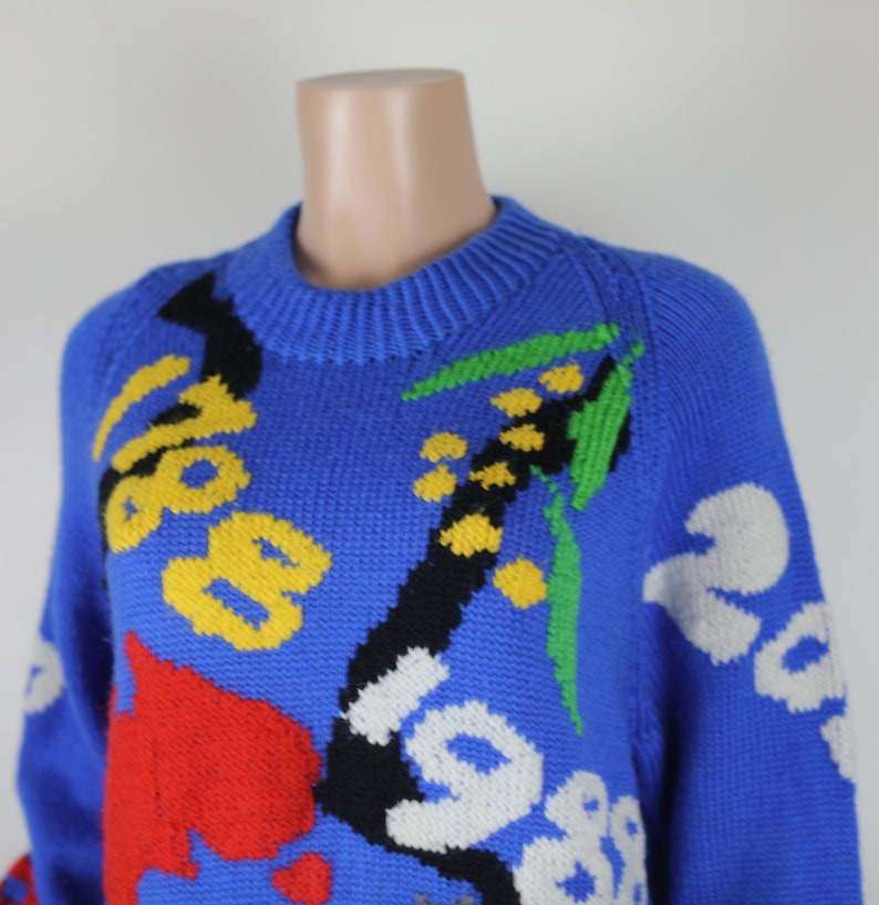 May include: A blue knitted jumper with a colourful abstract design. The design features yellow, red, green, black and white accents.