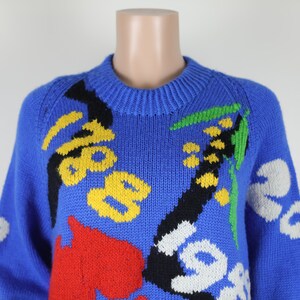 May include: A blue knitted jumper with a colourful abstract floral pattern. The jumper features yellow, red, black, white and green accents.