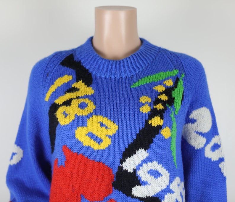 May include: A blue knitted jumper with a colourful abstract floral pattern. The jumper features yellow, red, black, white and green accents.