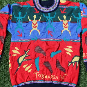 Pu&ograve; includere: Un colorato maglione lavorato a maglia con scollo tondo. Il maglione presenta una base rossa con varie forme astratte e la scritta "TORWABBA ART". La parte superiore ha sezioni blu e verdi con figure stilizzate e frecce.