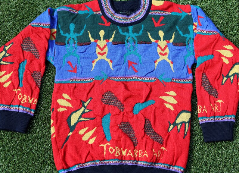Pu&ograve; includere: Un colorato maglione lavorato a maglia con scollo tondo. Il maglione presenta una base rossa con varie forme astratte e la scritta "TORWABBA ART". La parte superiore ha sezioni blu e verdi con figure stilizzate e frecce.