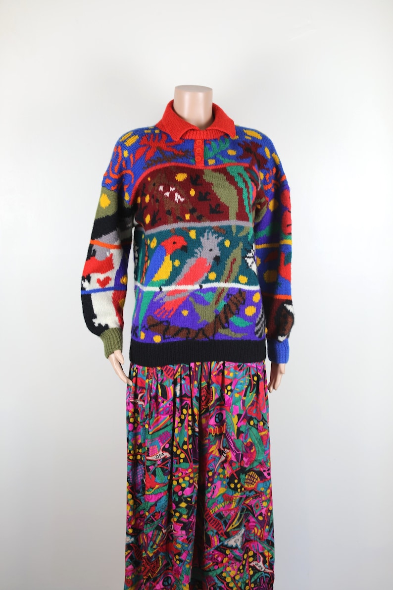 May include: A colorful knitted sweater with a multi-colored parrot design. The sweater has a red collar and a black band at the bottom.