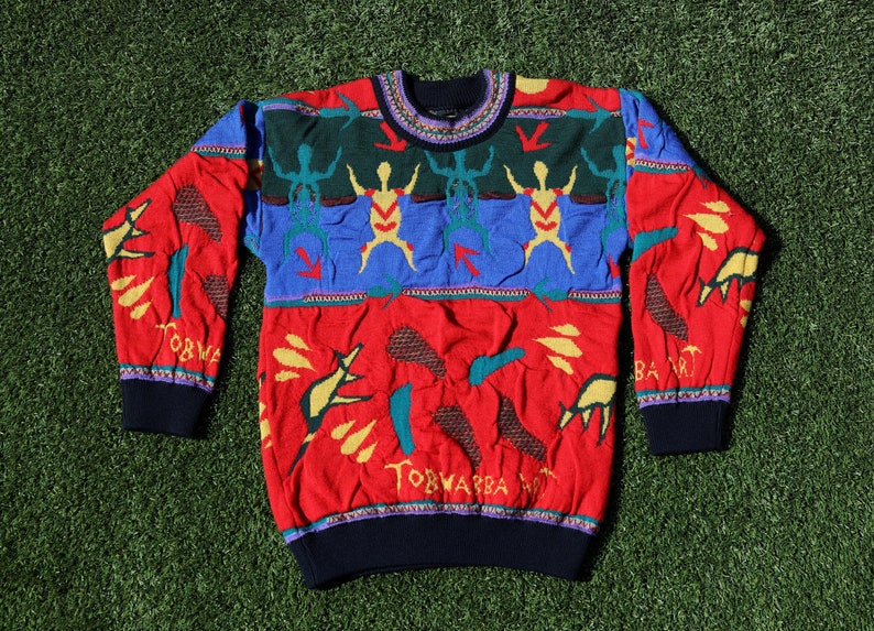Pu&ograve; includere: Un maglione colorato con girocollo e maniche lunghe. Il maglione presenta una base rossa con dettagli blu, verdi e gialli. Presenta un motivo di omini stilizzati, frecce e forme astratte. I polsini e lo scollo sono blu scuro.