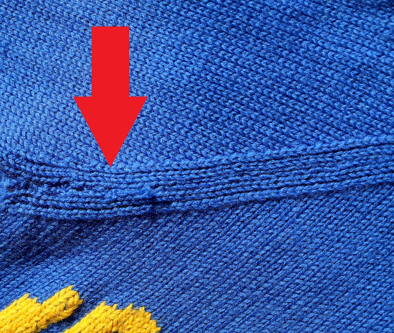 May include: Close-up of a blue knitted fabric with a seam running horizontally across the middle. The seam is highlighted by a red arrow.