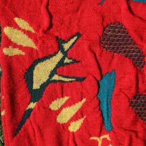 Pu&ograve; includere: Maglione rosso a maglia con un motivo ripetuto di forme astratte gialle e turchesi. Il maglione ha un aspetto strutturato e una visione ravvicinata del tessuto mostra i dettagli della maglia.