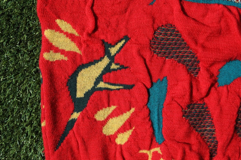 Pu&ograve; includere: Maglione rosso a maglia con un motivo ripetuto di forme astratte gialle e turchesi. Il maglione ha un aspetto strutturato e una visione ravvicinata del tessuto mostra i dettagli della maglia.