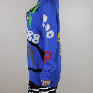 May include: A blue knitted jumper with a colourful, abstract design. The jumper has a crew neck and long sleeves.