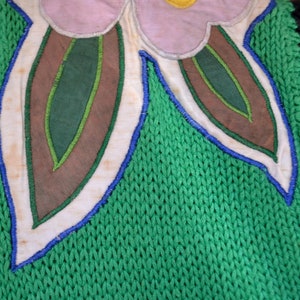 Puede incluir: Primer plano de una tela de punto verde con una aplicaci&oacute;n blanca, azul y marr&oacute;n de una flor con hojas.