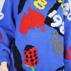 May include: A blue knitted jumper with a colourful, abstract design. The design features a red, yellow and black kangaroo, a red ladybug, and a red, yellow and black map of Australia.
