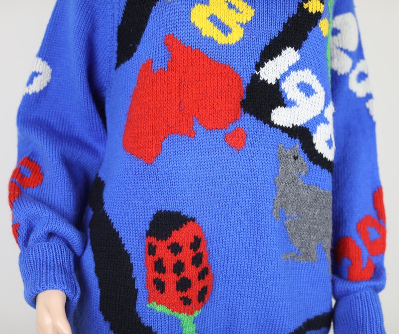 May include: A blue knitted jumper with a colourful, abstract design. The design features a red, yellow and black kangaroo, a red ladybug, and a red, yellow and black map of Australia.