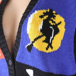 Pu&ograve; includere: Un gilet di maglia nero, blu, rosso, bianco e giallo con una strega che vola su una scopa davanti a un grafico di luna gialla.