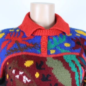 May include: A colorful knitted sweater with a red collar and three button closure. The sweater features a geometric pattern in shades of red, blue, green, yellow, and brown.