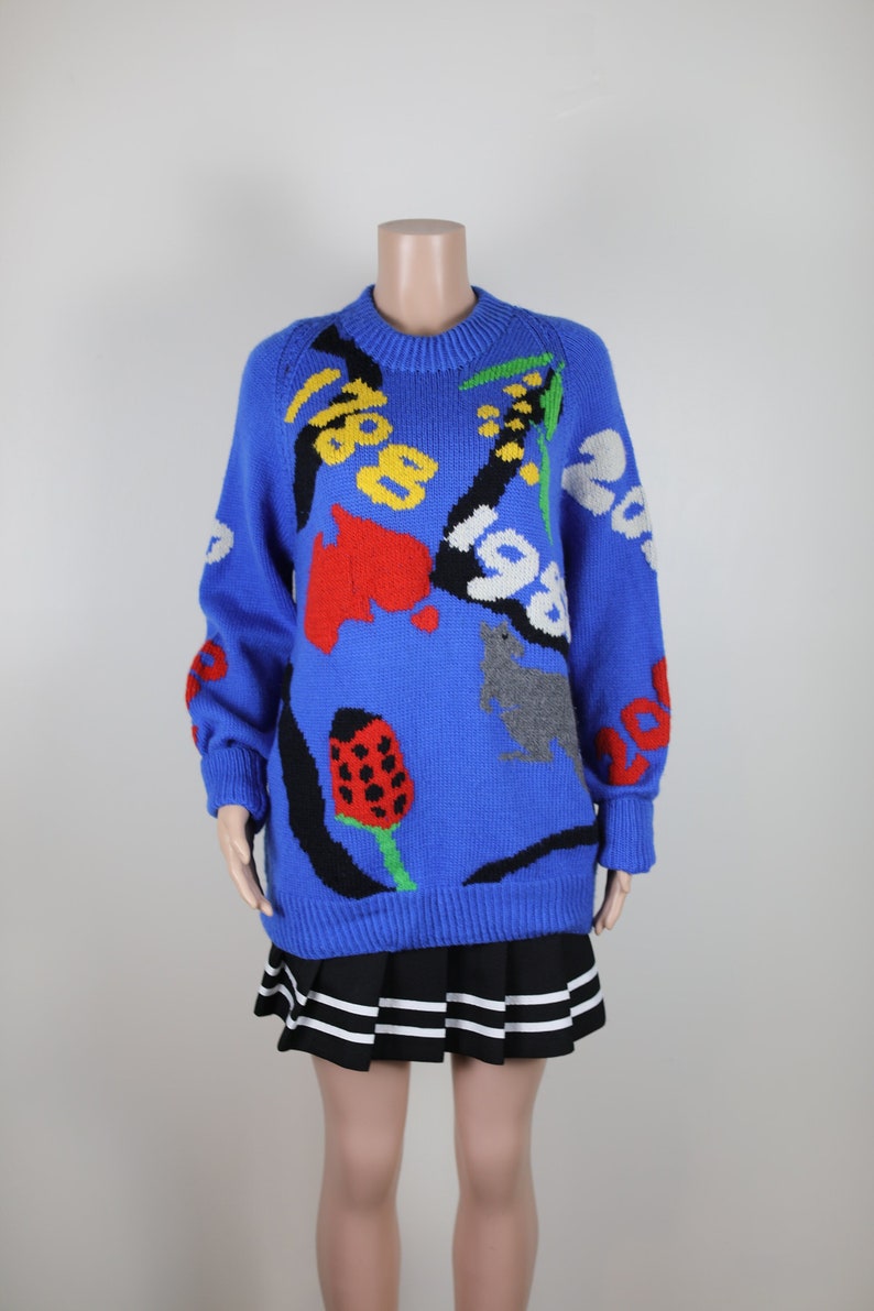 May include: A blue knitted jumper with a colourful abstract design featuring numbers, flowers, and animals. The jumper has long sleeves and a crew neck.