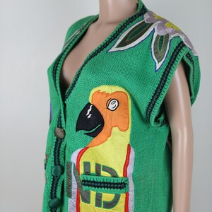 Puede incluir: Un chaleco de punto verde con un dise&ntilde;o de loro, que presenta un p&aacute;jaro amarillo, naranja y negro con un pico rojo. El chaleco tiene cuello en V y cierre de bot&oacute;n.