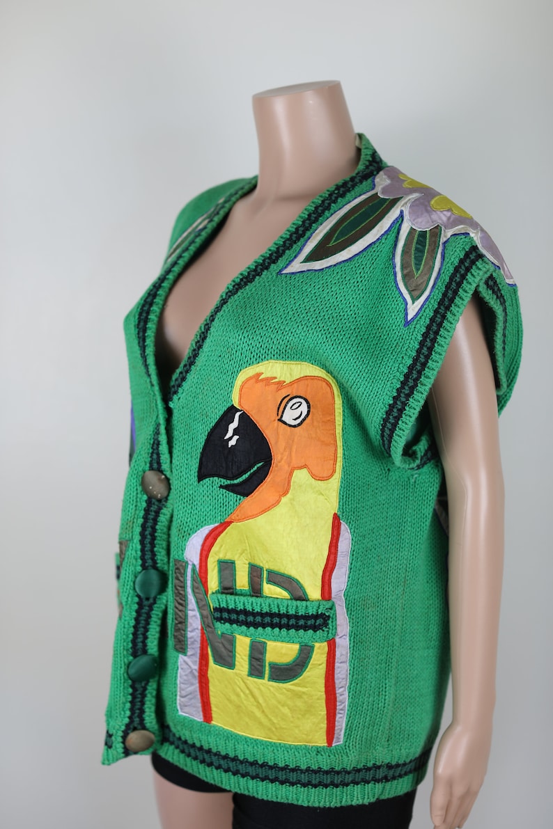 Puede incluir: Un chaleco de punto verde con un dise&ntilde;o de loro, que presenta un p&aacute;jaro amarillo, naranja y negro con un pico rojo. El chaleco tiene cuello en V y cierre de bot&oacute;n.