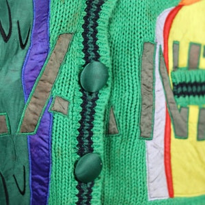 Puede incluir: C&aacute;rdigan de punto verde con parches bordados de colores y ribetes negros y verdes. El c&aacute;rdigan tiene cuatro botones grandes.