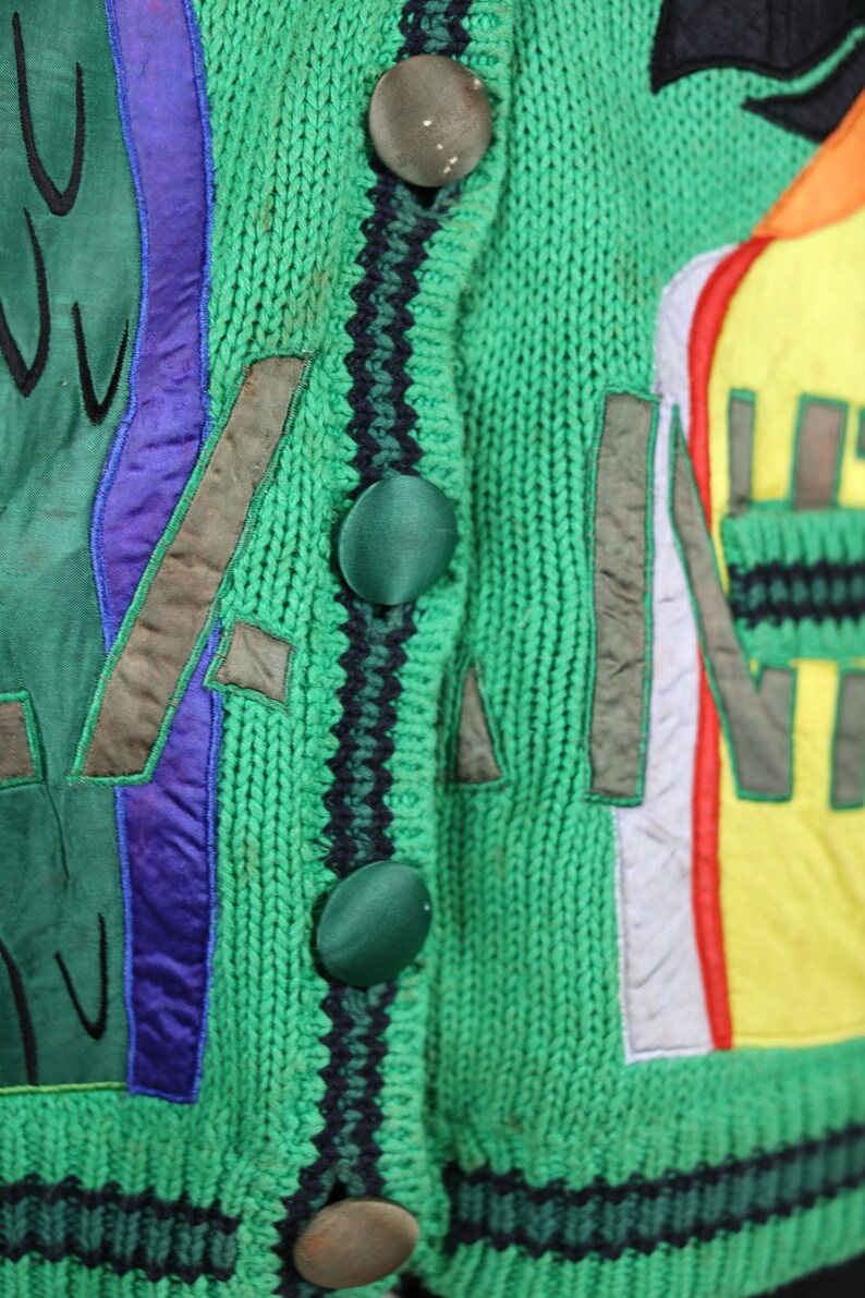 Puede incluir: C&aacute;rdigan de punto verde con parches bordados de colores y ribetes negros y verdes. El c&aacute;rdigan tiene cuatro botones grandes.