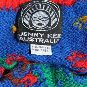 May include: A black and white tag with the text "JENNY KEE AUSTRALIA" and "SIZE TO FIT CM M BUST 85 CM". The tag is attached to a blue, red, green, and yellow knitted fabric.