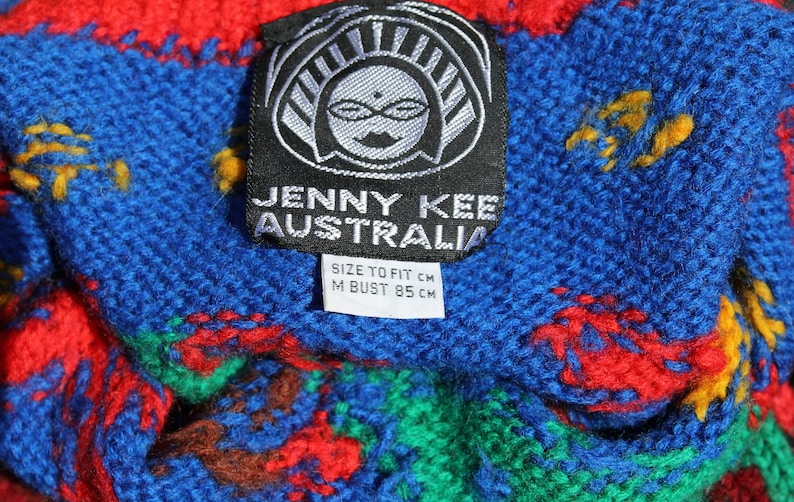 May include: A black and white tag with the text "JENNY KEE AUSTRALIA" and "SIZE TO FIT CM M BUST 85 CM". The tag is attached to a blue, red, green, and yellow knitted fabric.