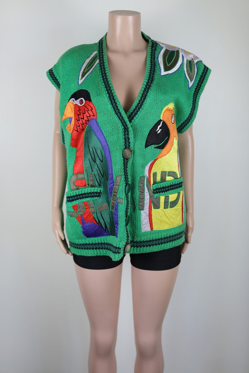 Puede incluir: Un chaleco de punto verde sin mangas con cierre de bot&oacute;n. El chaleco presenta loros bordados de colores y la palabra "Island" en la parte delantera.