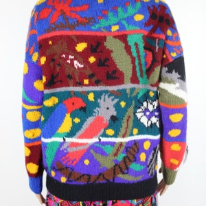 May include: A colorful knitted sweater with a multi-colored pattern of birds, flowers, and leaves. The sweater has a red collar and a black trim.