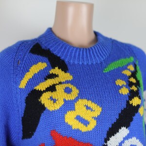 May include: A blue knitted jumper with a colourful abstract design. The jumper features yellow, black, red, green, and white accents.