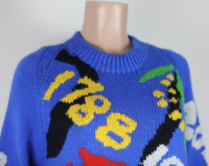 May include: A blue knitted jumper with a colourful abstract design. The jumper features yellow, black, red, green, and white accents.