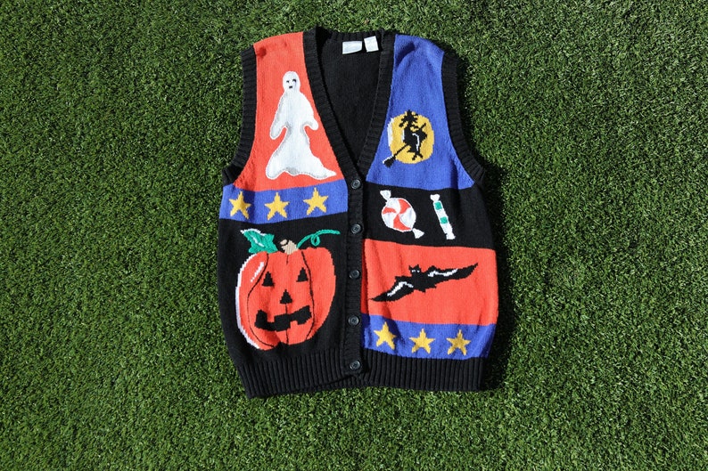 Pu&ograve; includere: Un gilet di maglia nero, rosso, blu e bianco a tema Halloween. Il gilet presenta un fantasma, un pipistrello, una zucca, bastoncini di zucchero e una strega su una scopa.