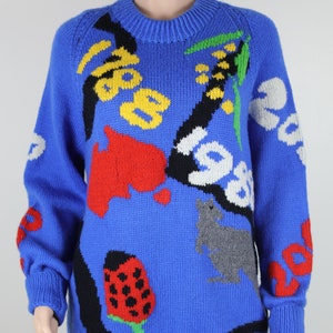 May include: A blue knitted jumper with a colourful abstract design. The design features a ladybug, a mushroom, a cat, and other abstract shapes.