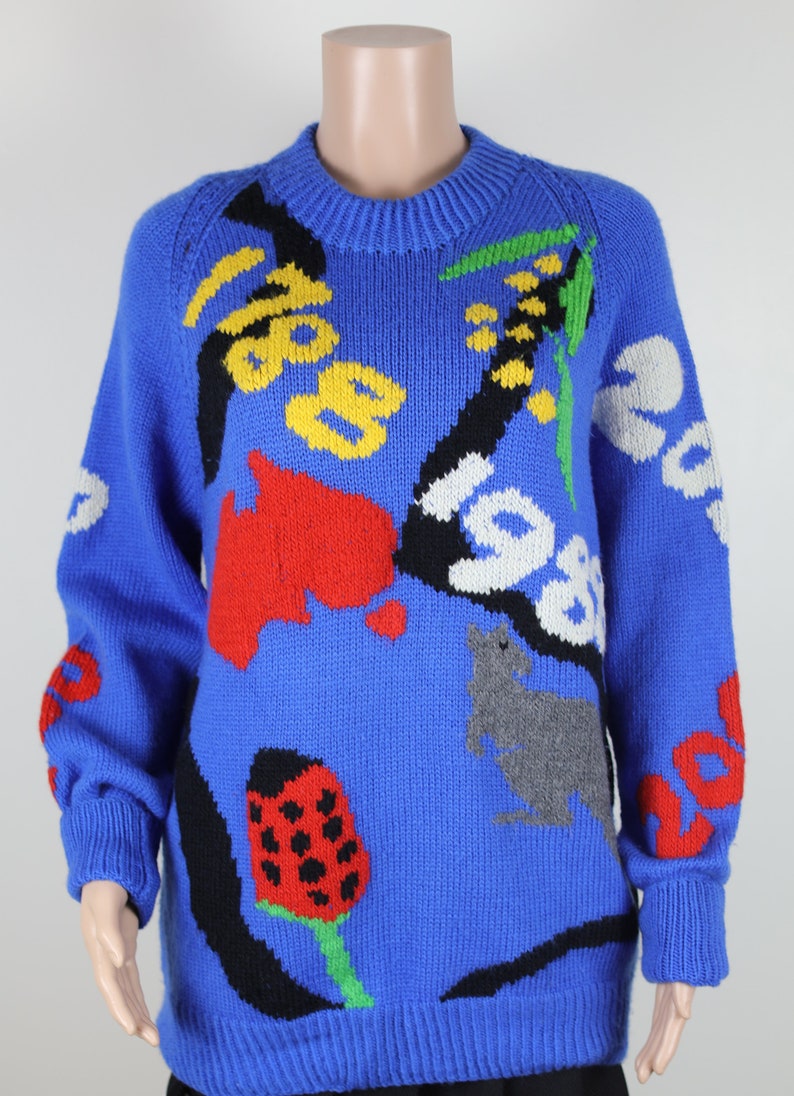 May include: A blue knitted jumper with a colourful abstract design. The design features a ladybug, a mushroom, a cat, and other abstract shapes.