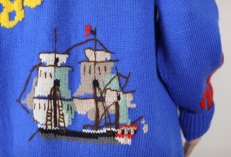 May include: A blue knitted jumper with a colourful embroidered ship design on the back. The ship has white, brown and green sails and a red flag.