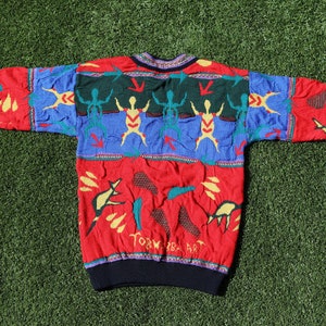 Pu&ograve; includere: Un maglione vintage colorato con scollo tondo. Il maglione presenta un motivo rosso, blu e verde con figure astratte e la scritta "TORK BABA ART". I polsini e lo scollo sono neri.