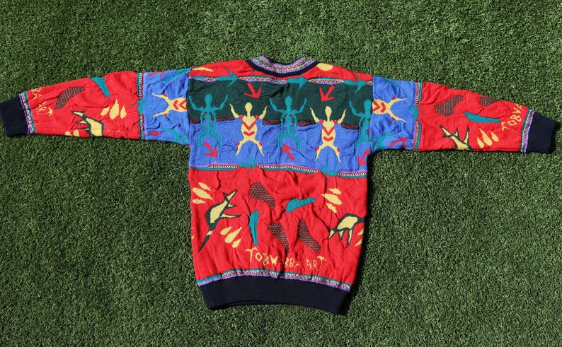 Pu&ograve; includere: Un maglione vintage colorato con scollo tondo. Il maglione presenta un motivo rosso, blu e verde con figure astratte e la scritta "TORK BABA ART". I polsini e lo scollo sono neri.