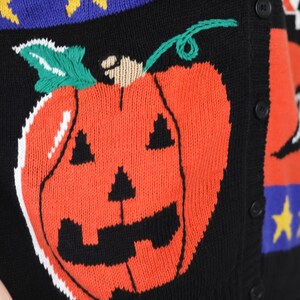 Pu&ograve; includere: Un cardigan di maglia nero con un design a patchwork colorato che presenta una grande zucca arancione con un viso di zucca intagliata. Il cardigan ha bottoni sul davanti.