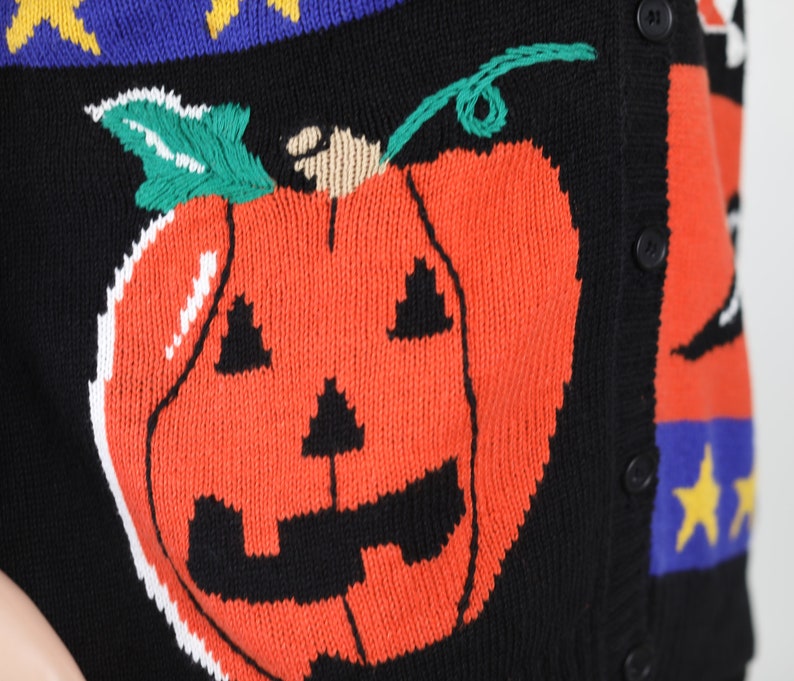 Pu&ograve; includere: Un cardigan di maglia nero con un design a patchwork colorato che presenta una grande zucca arancione con un viso di zucca intagliata. Il cardigan ha bottoni sul davanti.