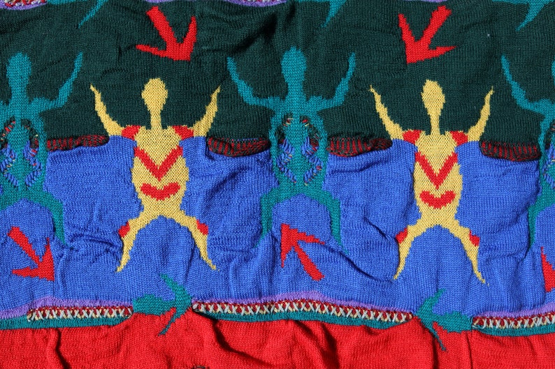 Pu&ograve; includere: Primo piano di un tessuto a maglia con un motivo ripetuto di figure stilizzate in giallo, verde acqua e rosso su uno sfondo blu e verde. Il design include frecce rosse e un bordo rosso e bianco.