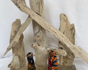 May include: A rustic nativity scene featuring two fabric-wrapped figures with dark skin tones, adorned in colorful African print fabrics. A seashell and driftwood form the backdrop, creating a unique and natural aesthetic.