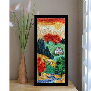 May include: A framed landscape art piece featuring a rural scene with a house, trees, and a river. The artwork uses a paper quilling technique, with vibrant colors and intricate details. The frame is black.