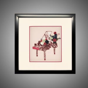 May include: A framed paper quilling art piece depicting a grand piano in shades of maroon and brown. The piano is adorned with a cluster of small, rolled paper flowers in red, white, and dark blue. The artwork is set within a black square frame.