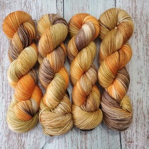 May include: Six skeins of hand-dyed yarn in shades of orange, brown, and yellow. The yarn is arranged in a row, with each skein showing a gradient of colors. The yarn is likely made of wool or a similar fiber, perfect for knitting or crocheting projects.