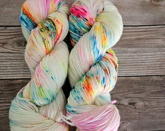 Puede incluir: Dos ovillos de hilo teñido a mano en una base blanca con salpicaduras de rosa brillante, naranja, amarillo, verde y azul.