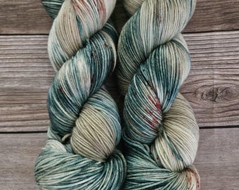 Puede incluir: Dos madejas de hilo teñido a mano con una paleta de colores variados en crema, verde salvia y azul cerceta profundo, acentuadas con sutiles motas de color rojo óxido a lo largo de las hebras retorcidas, creando un aspecto multitonal.