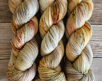 Puede incluir: Tres ovillos de hilo en una gama de colores beige, marrón y naranja. El hilo es una mezcla jaspeada con motas de color en todo el hilo.